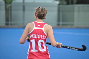 Mrs. Wright is pictures in her red hockey jersey during one of the world cup matches. she holds her hockey stick aloof and is pictured from behind. 